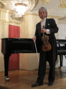 ヴァイオリンを手にピアノの前に立つ八嶋博人氏